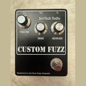 Černý efekt pedál s nápisem CUSTOM FUZZ a značkou DevTech Audio. Obsahuje tři stříbrné oválné knoflíky s popisky VOLUME, TONE a SUSTAIN. V horní část…