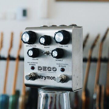 Kovový efekt pedál stříbrné barvy s nápisy DECO, tape saturation & doubletracker a strymon. Na horní části jsou tři černé otočné knoflíky s popisky S…