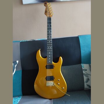 Zlatá elektrická kytara s tvarem superstrat a hnědým hmatníkem. Nástroj má jeden humbucker v pozici mostu, dva snímače typu single-coil a zlatý hardw…