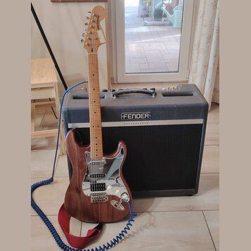 Elektrická kytara s hnědým dřevěným povrchem a bílým pickguardem stojí před černým kombem. Na hlavě kytary je nápis STRATOCASTER. Kombo má na přední…