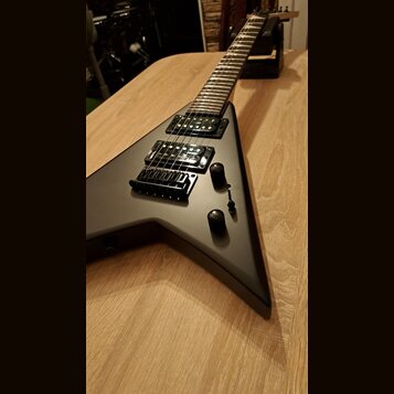 Matně černá elektrická kytara s tělem ve tvaru V položená na dřevěném stole. Nástroj má dva humbuckery, jeden potenciometr hlasitosti a jeden tónový…