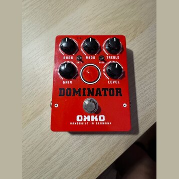 Červený efekt pedál s nápisy DOMINATOR a OHKO HANDBUILT IN GERMANY. Obsahuje pět černých otočných ovladačů s popisky BASS, MIDS, TREBLE, GRAIN a LEVE…