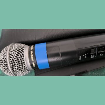 Detailní pohled na horní část černého bezdrátového mikrofonu s kovovou síťkou. Na těle je modrý ozdobný kroužek a nápis THE SHURE WIRELESS. Jsou vidě…