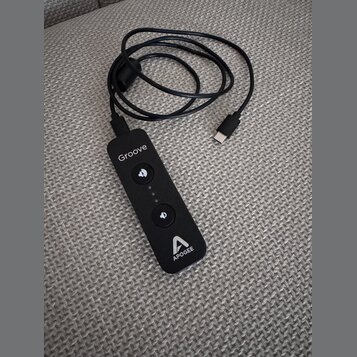 Černý přenosný audio rozhraní s připojeným kabelem USB-C. Na zařízení jsou bílé nápisy „Groove“ a „APOGEE“ spolu s logem značky. Uprostřed se nacháze…