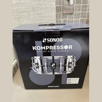 Černý papírový karton s nápisy SONOR, KOMPRESSOR SNARE DRUM SERIES a webovou adresou sonor.com. Na krabici je fotografie chromovaného malého bubnu s…