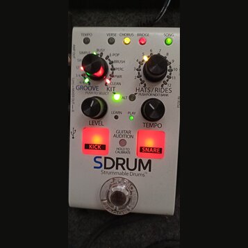 Bílý efekt pedál s nápisy GROOVE, KIT a HATS/RIDES. Na zařízení je vidět velké logo SDRUM a text Strummable Drums™. Obsahuje dva černé otočné knoflík…