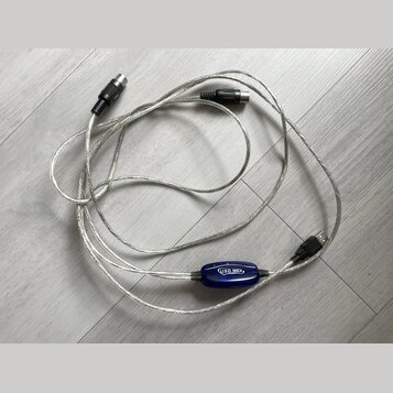 Stříbrný USB MIDI kabel s modrým plastovým převodníkem. Na převodníku je bílý nápis USB MIDI. Kabel má na koncích konektory USB a dva 5pinové DIN kon…