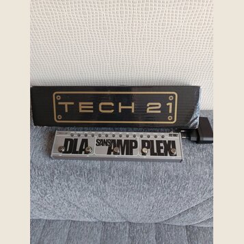 Na fotografii je kytarový pedál v hliníkovém šasi s nápisy TECH 21, SANSAMP PLEX! a DLA. Zařízení má tři stoupací spínače s popisy TAP, ON, ON a ON.…