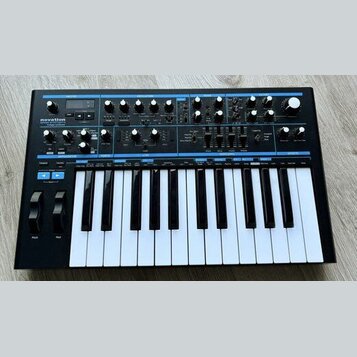 Černý syntezátor s klávesnicí a ovládacím panelem s modrými detaily. Na levé straně je bílý nápis novation a pod ním text Bass Station II analogue sy…