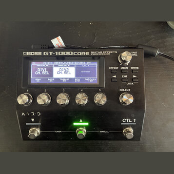 Černý kytarový procesor s nápisem BOSS GT-1000CORE GUITAR EFFECTS PROCESSOR. Zařízení má digitální displej, pět stříbrných otočných ovladačů v řadě,…