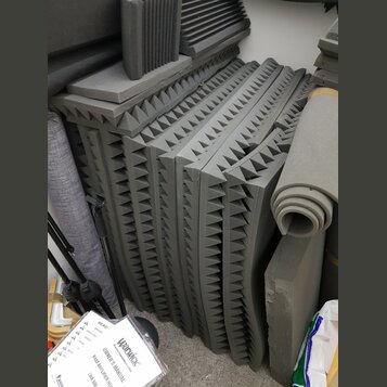 Svrhovatě naskládané šedé akustické pěny s pyramidovitým povrchem. Vedle nich je srolovaný šedý zvukový izolant a další kusy pěnového materiálu. V po…