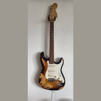 Elektrická kytara typu Stratocaster s černým lakem, který je na korpusu výrazně odloupaný a odhaluje světlé dřevo. Nástroj má bílý pickguard, tři sní…