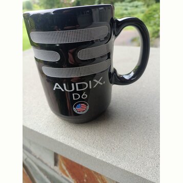 Černý keramický hrnek ve tvaru mikrofonu s vymalovanou stříbrnou mřížkou. Na povrchu je bílý nápis AUDIX. D6 a malá kulatá nálepka s vlajkou USA a ná…