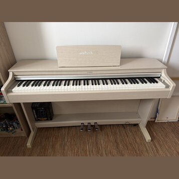 Digitální piano v krémové barvě s povrchem imitujícím světlé dřevo. Nad klávesnicí je umístěn nápis YAMAHA a na hudebním stojanu je vidět vybledlé bí…