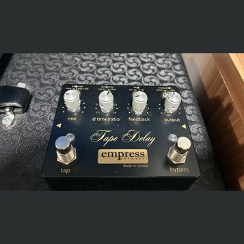 Černý efekt pedál s nápisem Tape Delay a logem empress effects. Na horní části jsou čtyři stříbrné oválné ovladače s popisky mix, d time|ratio, feedb…