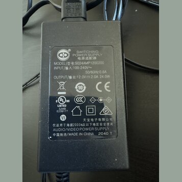 Černý plastový síťový adaptér s nápisem SWITCHING POWER SUPPLY a AUDIO/VIDEO POWER SUPPLY. Na štítku je uveden model S024AMP1200200, vstup INPUT 100-…