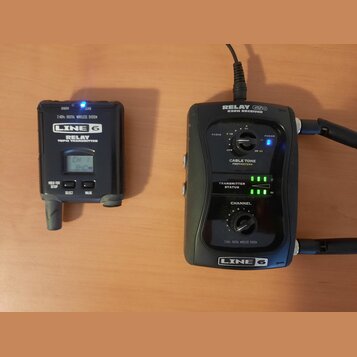 Na fotografii jsou dva černé přístroje značky LINE 6. Vlevo je vysílač s nápisy RELAY G50 TRANSMITTER a 2.4GHz DIGITAL WIRELESS SYSTEM, vybavený malý…