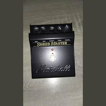 Černý kytarový efekt pedál s nápisy Gain, Bass, Contour, Treble a Volume. Uprostřed je velký bílý nápis SHRED MASTER a ve spodní části vy embossed lo…