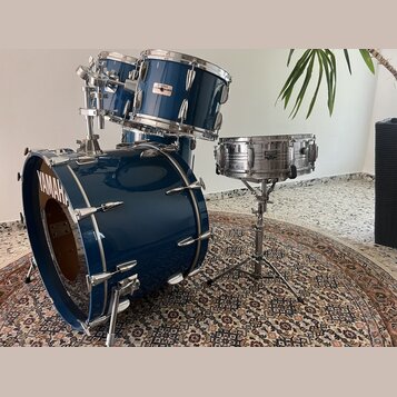 Modrá bicí souprava s kovovým hardwarem. Na basovém bubně je bílý nápis YAMAHA, na malém bubnu je viditelné logo YAMAHA. Na jednom z tomů je nalepený…