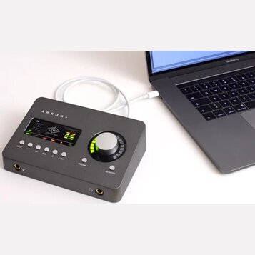 Šedý audio rozhraní s nápisem ARROW připojený bílým kabelem k notebooku MacBook Pro. Zařízení má digitální displej s logem UNIVERSAL AUDIO, ovládací…