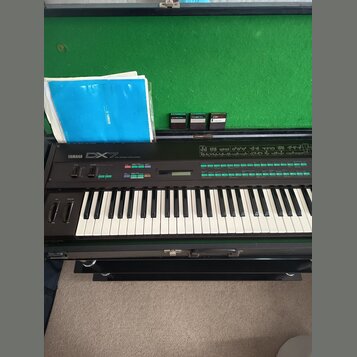 Černý digitální syntezátor s klávesnicí, na kterém je nápis YAMAHA DX7. Nástroj je umístěn v otevřeném transportním kufru s zeleným vnitřkem, ve kter…