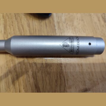 Detail stříbrného kovového těla mikrofonu. Na povrchu je vyryto logo v trojúhelníku a nápisy BEHRINGER a ECM 8000. V pravé části je vidět malý kulatý…