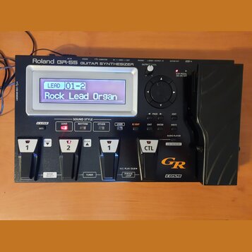Černý kytarový syntetizátor s nápisem Roland GR-55 GUITAR SYNTHESIZER. Zařízení má digitální displej, na kterém je text LEAD 01-2 Rock Lead Organ, ně…