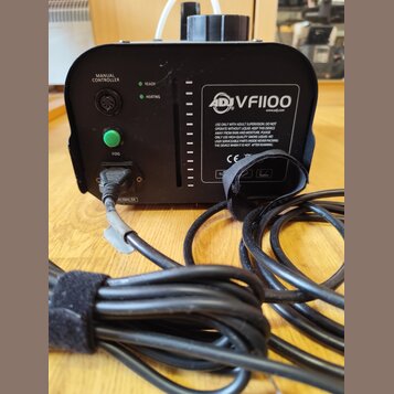 Černý kouřový stroj s nápisem ADJ VF1100 a webovou adresou www.adj.com. Na předním panelu jsou kontrolky READY a HEATING, zelené tlačítko FOG a nápis…