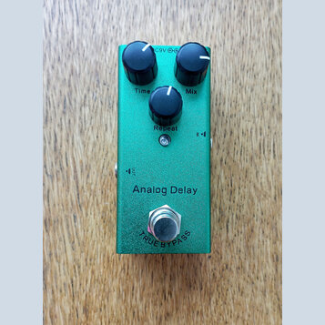 Zelený efekt pedál Analog Delay s kovovým povrchem. Na horní straně jsou tři černé oválné ovladače s popisky Time, Mix a Repeat. V dolní části je sti…