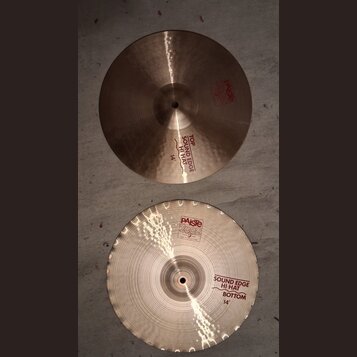 Dvě mosazné činely typu hi-hat o průměru 14". Horní činel nese červené nápisy „PAISTE 2002“ a „TOP SOUND EDGE HI-HAT 14"“. Spodní činel má v horní čá…