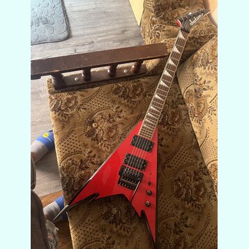 Červená elektrická kytara s tělem ve tvaru V a černými hranami. Na hlavě hmatníku je bílé logo Jackson. Kytara má dva černý humbuckery, černý most s…