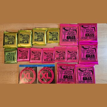 Sada různých balení kytarových a basových strun značky Ernie Ball. Horní řada obsahuje čtyři žluté balení s nápisem REGULAR Slinky a dvě růžové balen…