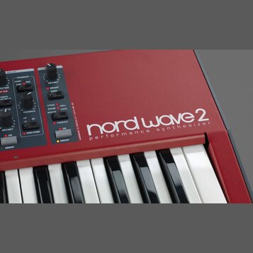 Detailní pohled na červený syntetizátor s klávesnicí. Na šasi je bílý nápis nord wave 2 a pod ním text performance synthesizer. V levé části jsou vid…