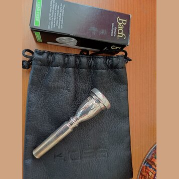 Stříbrný kovový moutur položený na černém koženém vakové pouzdře. Vedle nich je černá krabička s nápisy Bach Genuine Mouthpiece a LEAD FREE. Na moutu…