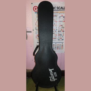 Černý tvrdý kufýr na kytaru s texturovaným povrchem. Ve spodní části je bílý nápis Gibson USA. Kufýr má připevněnou černou plastovou rukojeť a kovové…