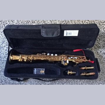 Zlatý saxofon v černém polstrovaném kufru. V horní části kufru je bílá nálepka s nápisem SAXOFON SERVIS JANA, QR kódem a kontaktními údaji. Vedle nás…