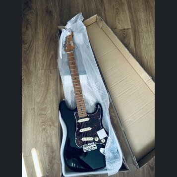 Černá elektrická kytara s tmavě červeným šarlatanem (pickguard) v kartonové krabici s ochrannou bublinkovou fólií. Na hlavě hmatníku je bílý nápis "S…