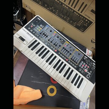 Bílo-šedý syntetizátor s klávesnicí a ovládacím panelem s mnoha posuvníky a tlačítky. Na horní části je nápis Roland GAIA SYNTHESIZER SH-01. Povrch p…