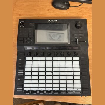 Černý MIDI kontrolér značky AKAI PROFESSIONAL s velkým displejem v horní části. Zařízení obsahuje mřížku 64 bílých podsvícených padů, několik ovládac…