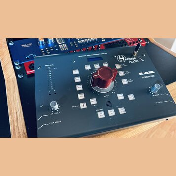 Černý monitorovací kontrolér s nápisy Heritage Audio a R.A.M. SYSTEM 2000. Uprostřed se nachází velký červený otočný knoflík, vedle něj jsou tlačítka…