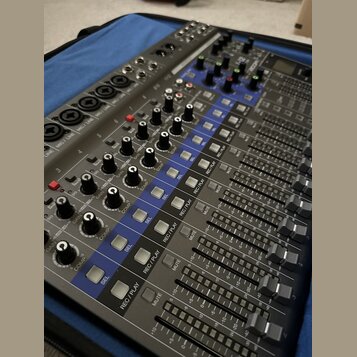 Šedý mixážní pult s modrými prvky a řadou posuvníků. Na panelu jsou viditelné nápisy jako GAIN, COMP, SEL, REC/PLAY a MUTE. Zařízení obsahuje početné…