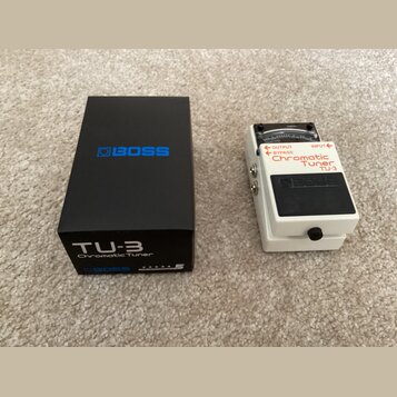 Bílo-černý ladicí pedál s nápisy OUTPUT, BYPASS, INPUT, Chromatic Tuner a TU-3. Na černé části je umístěno logo BOSS. Vedle pedálu je černá krabice s…