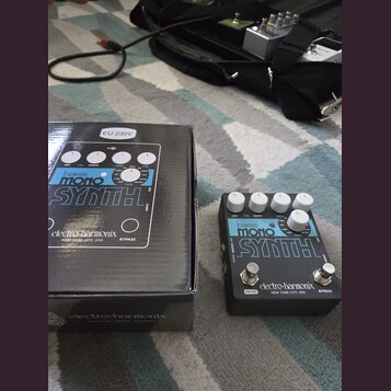 Na fotografii je efekt pedál Bass Mono Synth značky electro-harmonix v černé barvě s bílými nápisy. Vedle pedálu je umístěna jeho originální krabice…