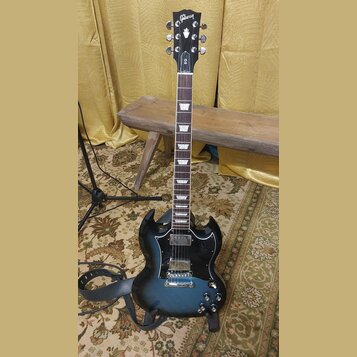 Černá elektrická kytara typu SG s modrým přechodem (sunburst) na korpusu. Na hlavě je bílý nápis Gibson a označení modelu SG. Nástroj má hnedý hmatní…