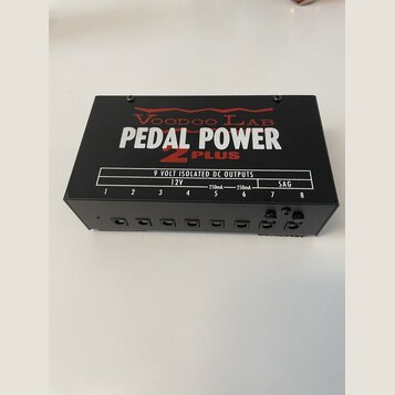 Černý napájecí zdroj pro efektové pedály s nápisy Voodoo Lab, PEDAL POWER 2 PLUS a tabulkou specifikací 9 VOLT ISOLATED DC OUTPUTS. Na přední straně…