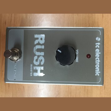 Stříbrný efekt pedál s nápisy RUSH a BOOSTER. Na pravé straně je logo tc electronic, vprostřed černý ovládací knoflík s popiskem VOLUME a červená kon…