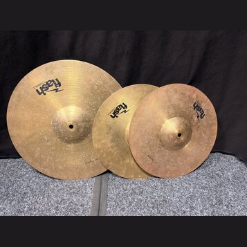 Tři mosazné činely různých velikostí s viditelným povrchovým opotřebením a šmouhama. Každý činel nese logo „flash CYMBALS“. Na okraji největšího čine…