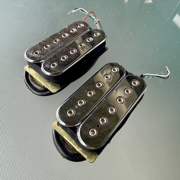 Dva elektrické kytarové snímače typu humbucker s chromovaným krytem a viditelnými poloslníky. Snímače mají připojené elektrické vodiče a jsou zespodu…
