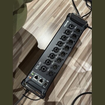 Černý digitální stagebox s kovovým šasi a madlymi po stranách. Na horní části je nápis Midas a řada konektorů XLR označených jako OUT 1 až OUT 8. V d…