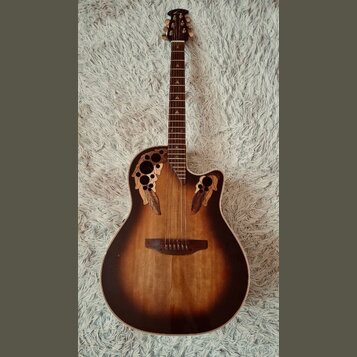 Akustická kytara s vyřezávanými otvory v horní desce místo klasického rozetového otvoru. Nástroj má tmavě hnědý přechodový povrch (sunburst) a hmatní…